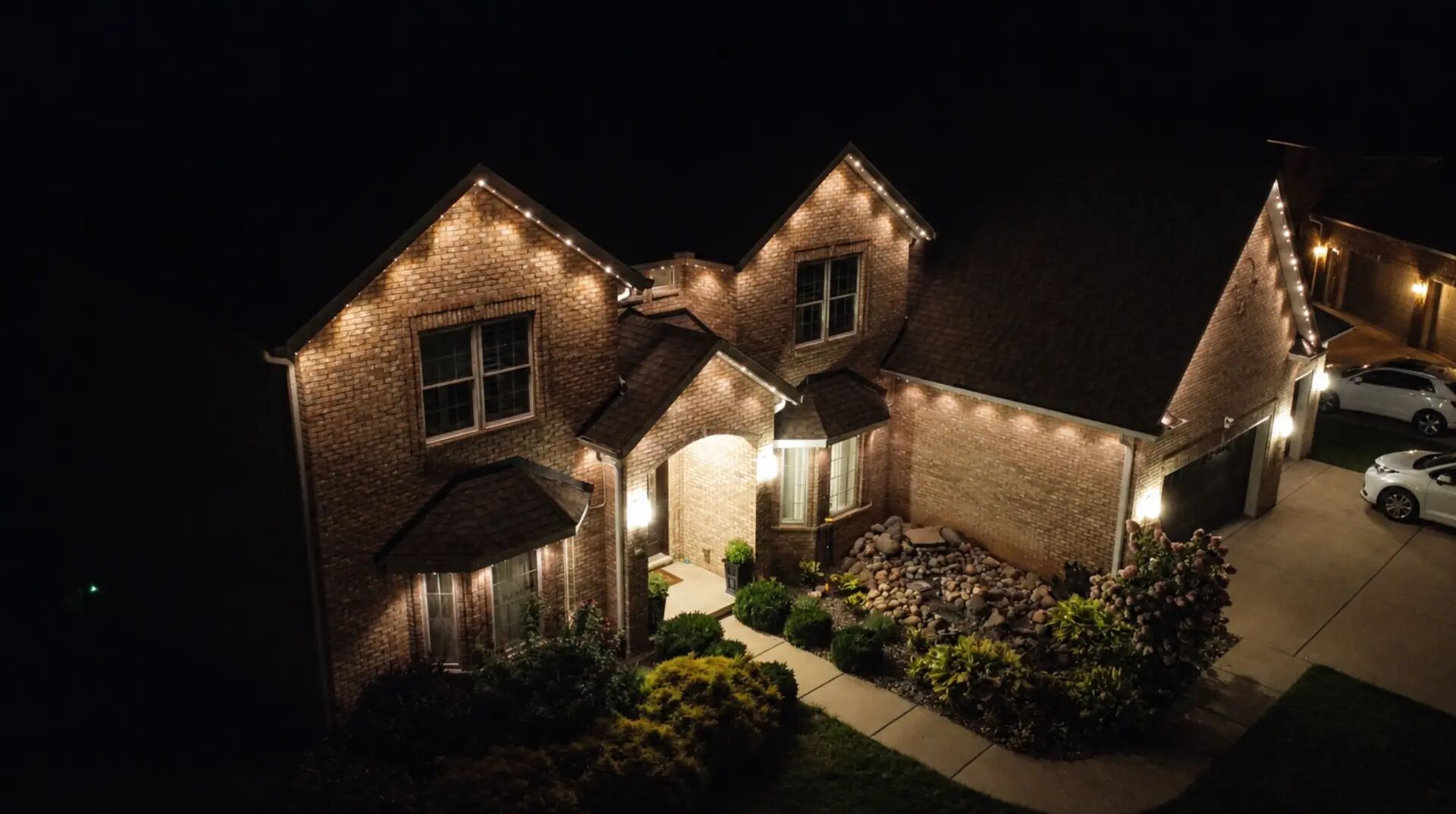 Aerial night view of a brick home with warm white lights along each gable and roofline
