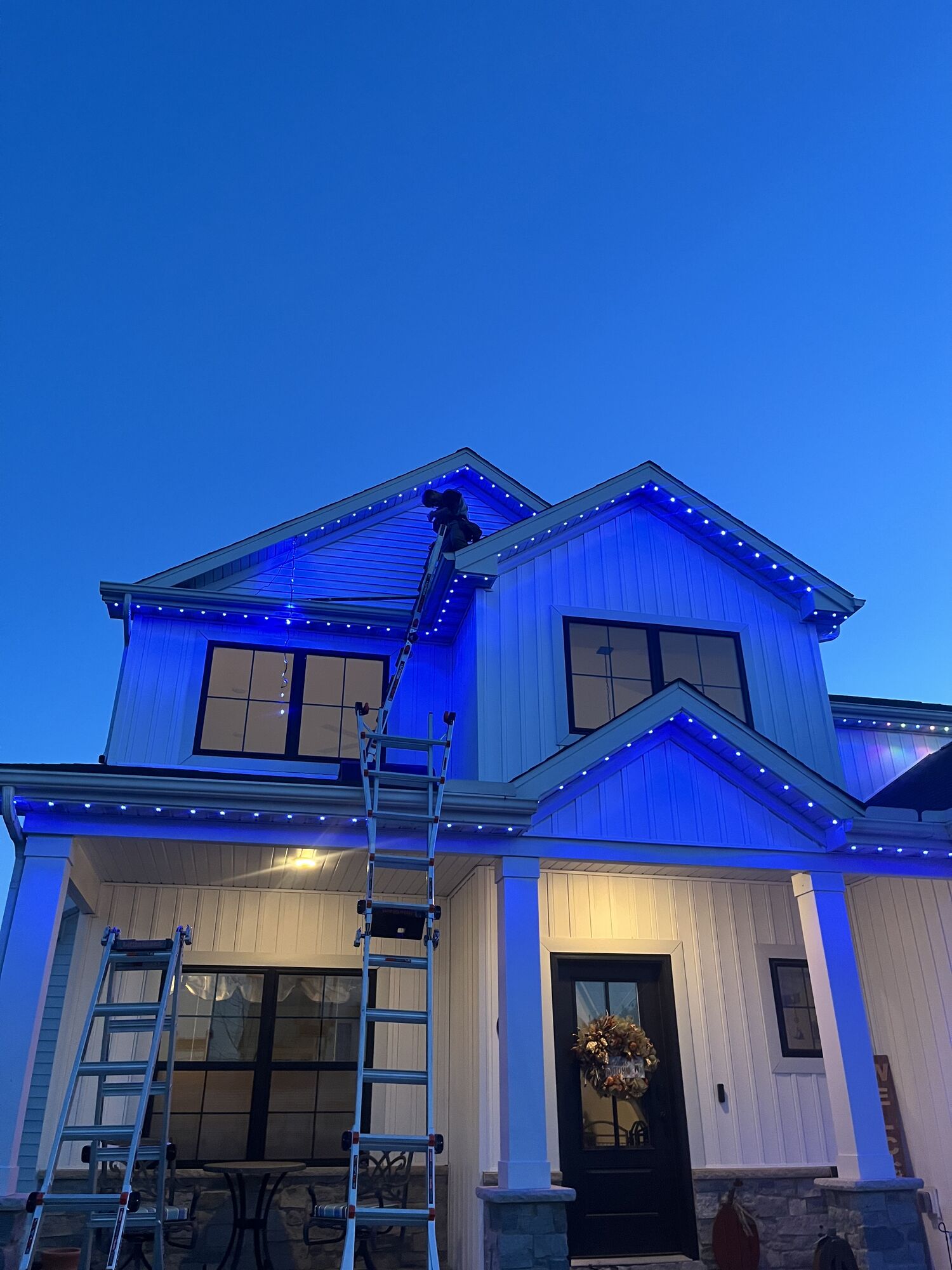 White farmhouse-style home at dusk with blue lighting along its gables, ladders still at the porch