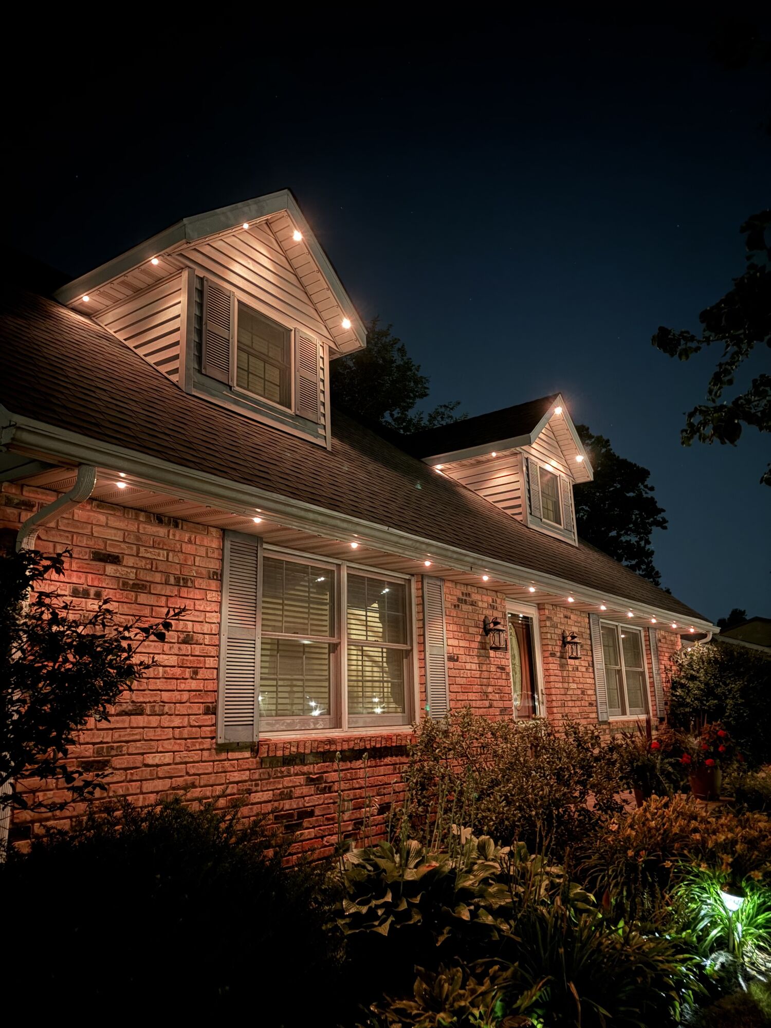 Brick home with warm white lights along the eaves and dormer peaks at night