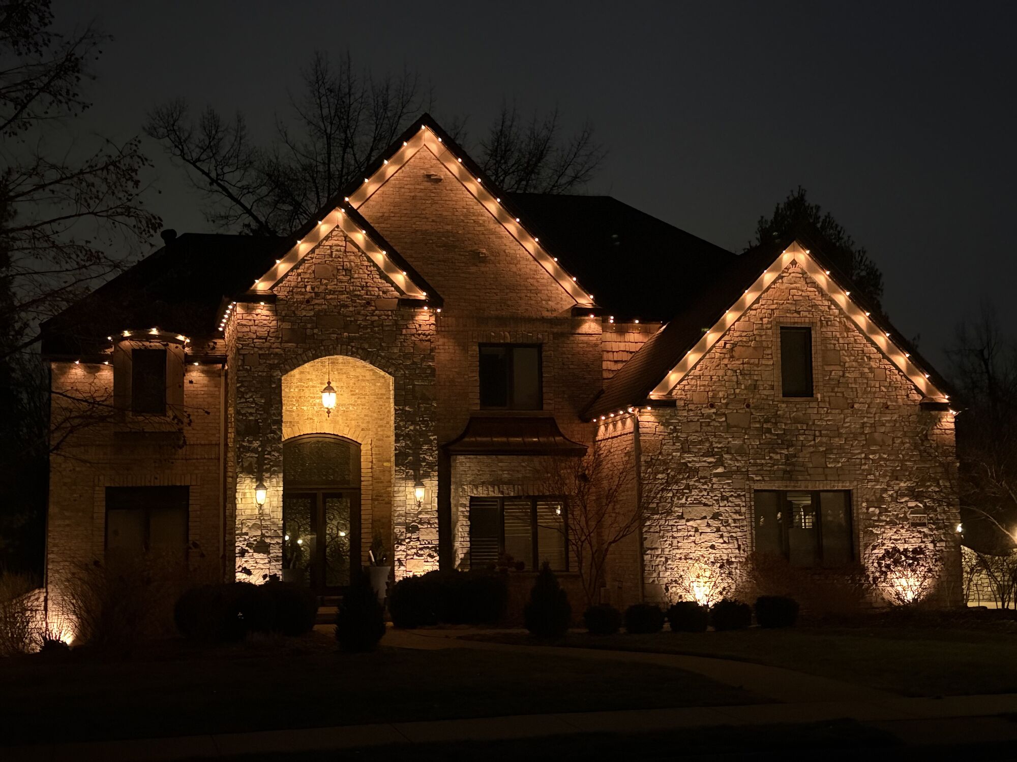 Stone home with warm white lights tracing multiple gables above the entryway