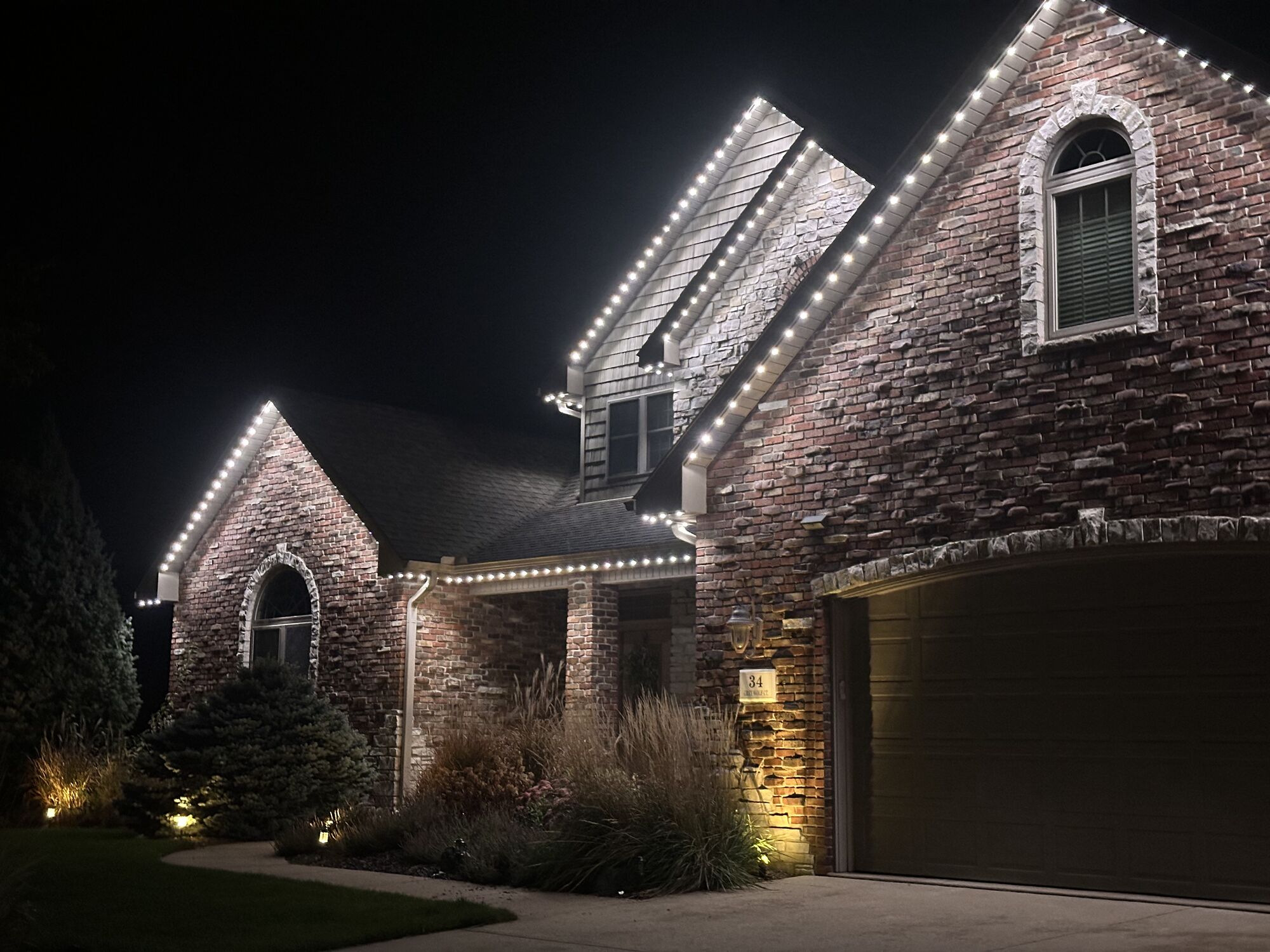 Brick home with crisp white lights tracing its steep rooflines and arched window gable