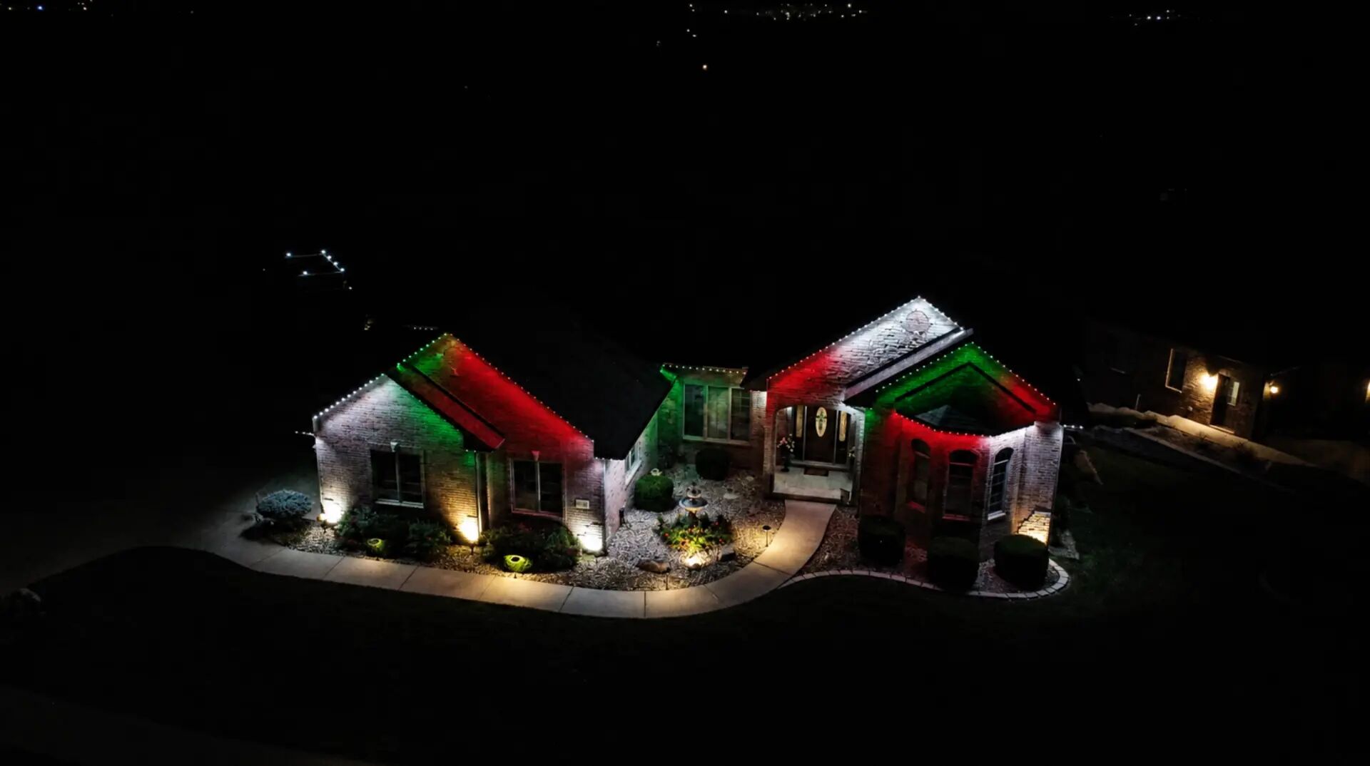 Aerial night view of a home with red, green and white lighting tracing its rooflines and landscape lights along the path