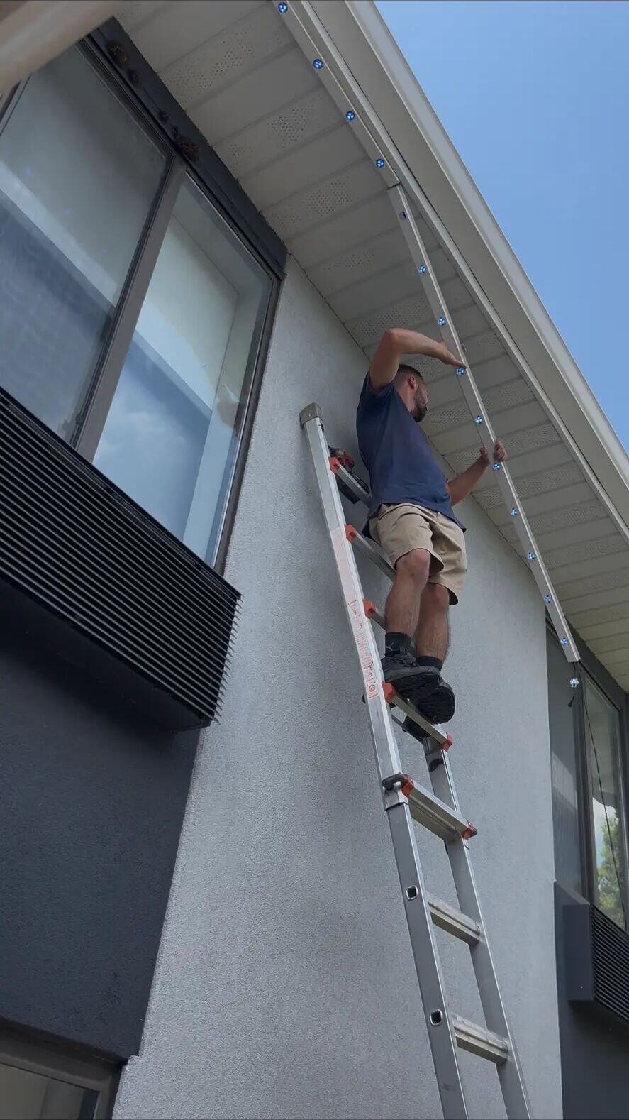 Installer on a ladder fitting lighting along the eaves of a home in daylight